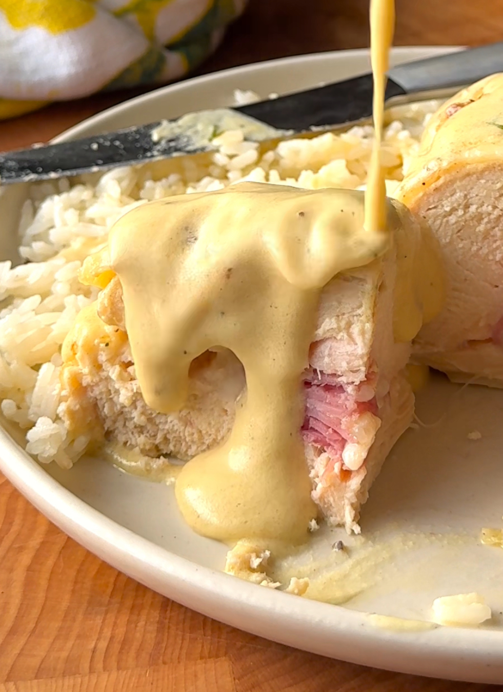 a slice of chicken stuffed with ham and cheese with sauce being poured over it, beside white rice on a plate